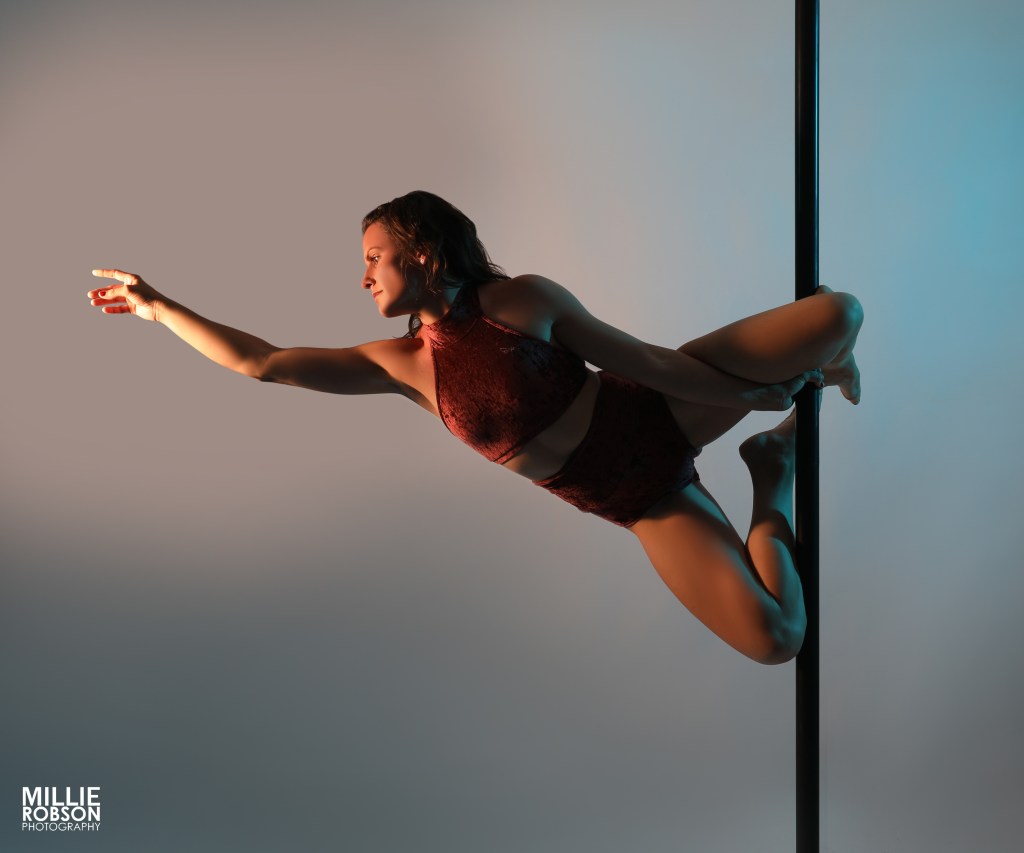 Eine Sportlerin führt eine anspruchsvolle Pose am Pole durch und streckt ihren Arm nach vorne, während sie an einer Pole befestigt ist. Der Hintergrund ist in sanften Farben gehalten.